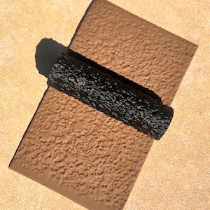 May include: A black textured roller sits on top of a brown textured surface. The roller is used for applying paint or other coatings.