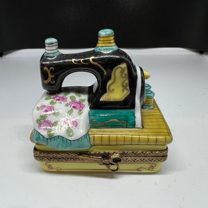 Könnte beinhalten: Dekorative Nähmaschinenfigur. Die schwarze Maschine hat goldene Akzente und sitzt auf einem gelben Sockel. Der Sockel hat ein Blumenmuster und eine Schere. Die Garnrollen sind blau.