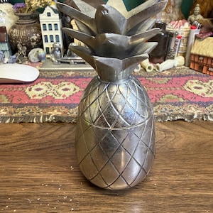 Puede incluir: Un recipiente de metal plateado con forma de piña y tapa extraíble. El cuerpo presenta una superficie texturizada con un patrón de diamantes, y la parte superior está adornada con hojas puntiagudas y superpuestas que forman la corona.
