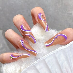 May include: Set of five long, nude-colored false nails with a white and purple abstract design. Each nail has a small, clear rhinestone accent.