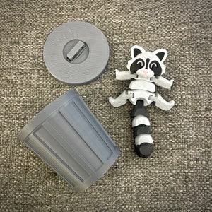 May include: A gray plastic trash can with a lid and a gray plastic raccoon toy with a black and white tail.