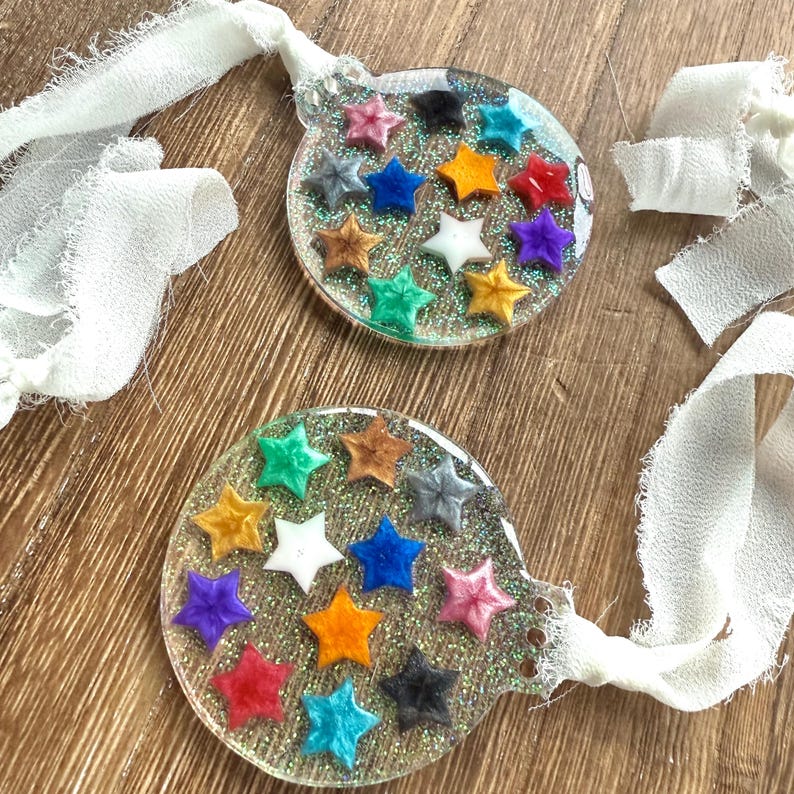 May include: Two clear, round ornaments with glitter and colorful star-shaped embellishments. Each ornament has a white ribbon for hanging. The stars are various colors, including red, blue, yellow, and green. The ornaments are on a wooden surface.