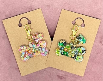 May include: Two dog bone-shaped pet ID tags with gold keychains. The tag on the left is pink and white with the name "PENNY" spelled out in colorful letters. The tag on the right is green and black with the name "TUCKER" spelled out in colorful letters.
