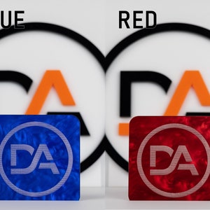 May include: Two square pieces of acrylic with a marbled pattern. The left piece is blue with a white "DA" logo inside a circle. The right piece is red with a white "DA" logo inside a circle. The text "BLUE" is above the left piece and "RED" is above the right piece.