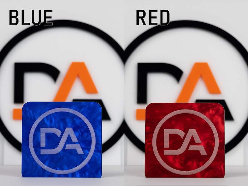 May include: Two square pieces of acrylic with a marbled pattern. The left piece is blue with a white "DA" logo inside a circle. The right piece is red with a white "DA" logo inside a circle. The text "BLUE" is above the left piece and "RED" is above the right piece.