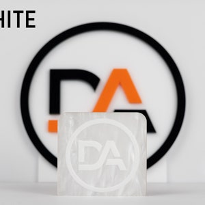 May include: White square tile with a white circle and the letters "DA" inside. The tile is sitting in front of a black circle with an orange "A" and a black "D" inside.