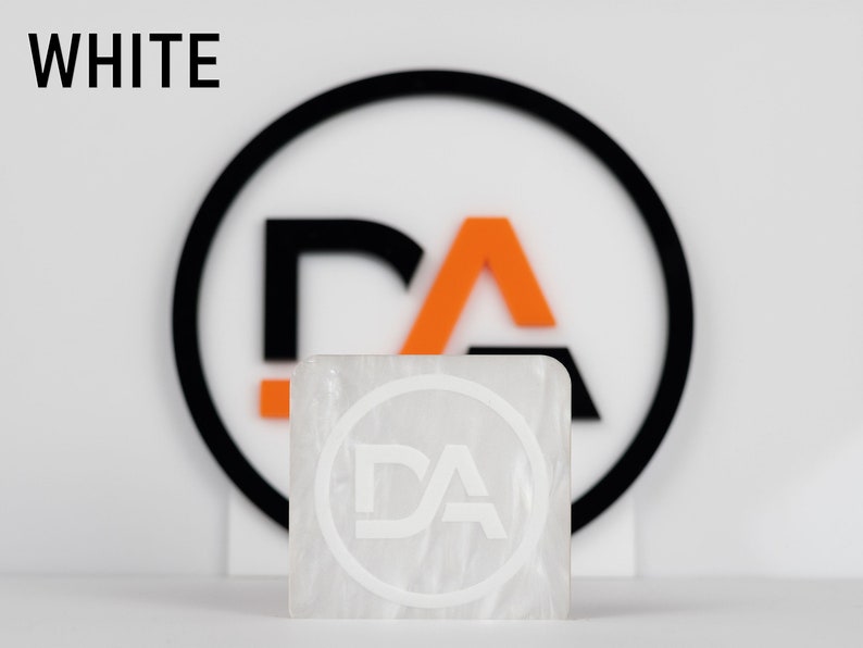 May include: White square tile with a white circle and the letters "DA" inside. The tile is sitting in front of a black circle with an orange "A" and a black "D" inside.