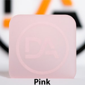 May include: A square, pale pink object with rounded corners. It features a subtle circular design with the letters "DA". The word "Pink" is printed in black at the bottom of the image.