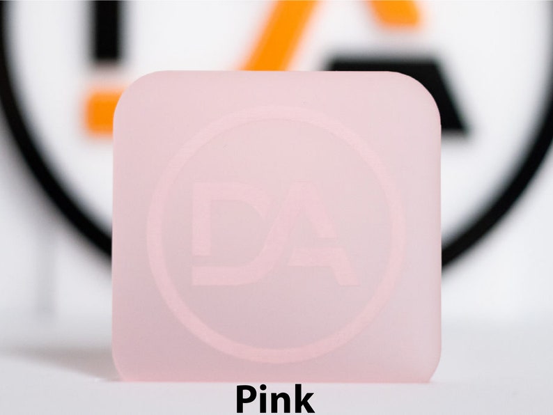 May include: A square, pale pink object with rounded corners. It features a subtle circular design with the letters "DA". The word "Pink" is printed in black at the bottom of the image.