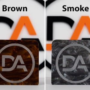 May include: Two square pieces of marbled plastic with a silver circle and the letters "DA" inside. The left piece is brown and the right piece is gray.