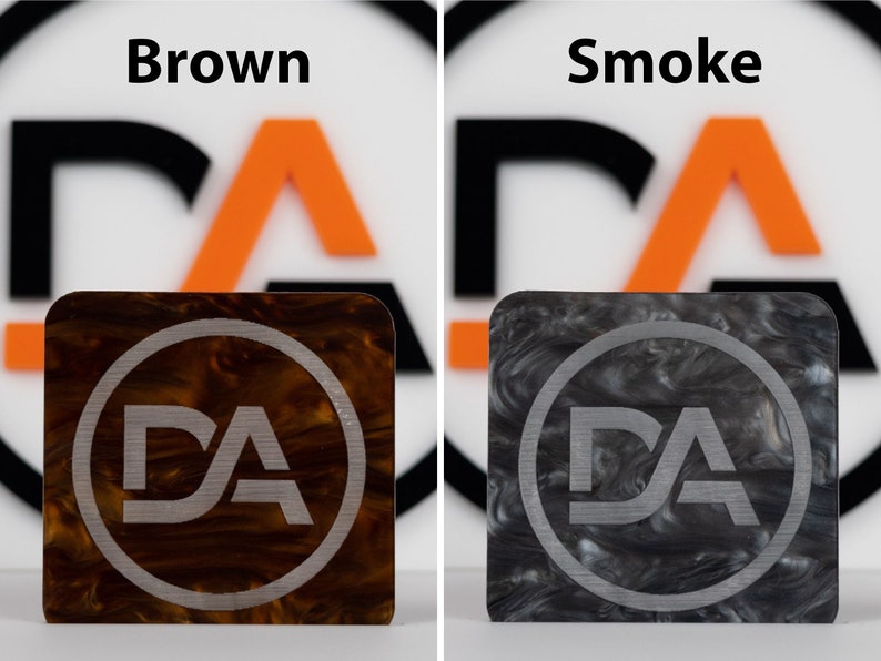 May include: Two square pieces of marbled plastic with a silver circle and the letters "DA" inside. The left piece is brown and the right piece is gray.