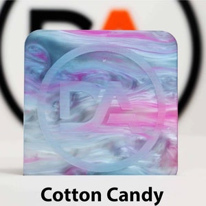 May include: A square, translucent, blue, pink, and white marbled resin tile with a white circle and the letters "DA" inside the circle.