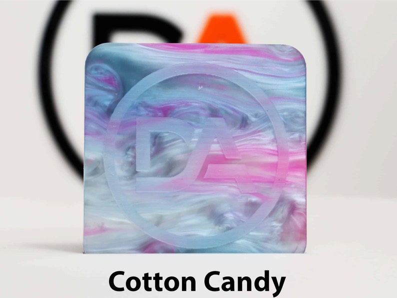 May include: A square, translucent, blue, pink, and white marbled resin tile with a white circle and the letters "DA" inside the circle.