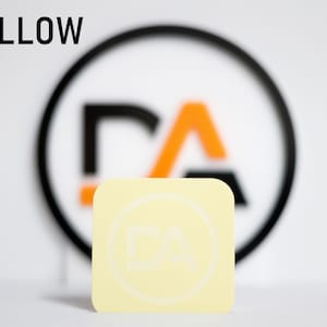 May include: A yellow square sticker with a white circle and the letters "DA" inside. The sticker is in front of a blurred background with a black circle and the letters "DA" inside.