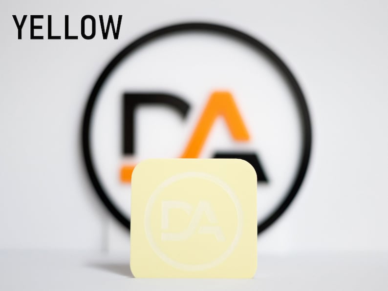 May include: A yellow square sticker with a white circle and the letters "DA" inside. The sticker is in front of a blurred background with a black circle and the letters "DA" inside.