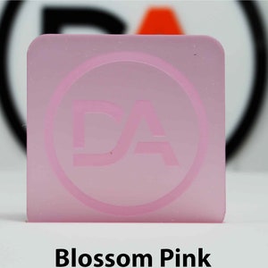 May include: A square, translucent, blossom pink acrylic piece with rounded corners. A circular logo with the letters "DA" is etched into the surface. The text "Blossom Pink" is printed in a bold, black font at the bottom of the image.