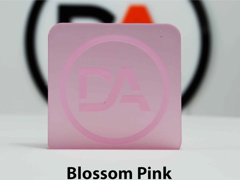 May include: A square, translucent, blossom pink acrylic piece with rounded corners. A circular logo with the letters "DA" is etched into the surface. The text "Blossom Pink" is printed in a bold, black font at the bottom of the image.