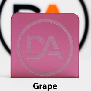 May include: A square, grape-coloured acrylic piece with rounded corners. A circular design with the letters "DA" is etched into the surface. The word "Grape" is printed in black at the bottom of the image.