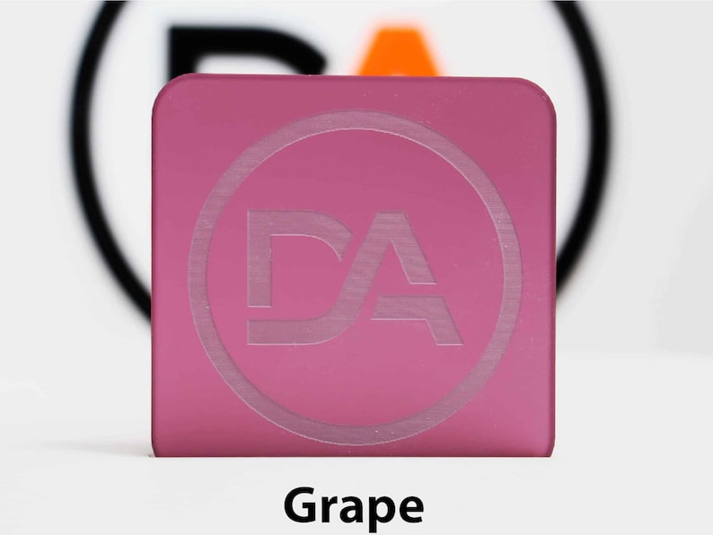 May include: A square, grape-coloured acrylic piece with rounded corners. A circular design with the letters "DA" is etched into the surface. The word "Grape" is printed in black at the bottom of the image.