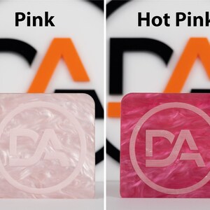 May include: Two square samples of acrylic plastic, one pink and one hot pink, with a white "DA" logo in a circle in the center of each.