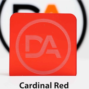 May include: A square, cardinal red object with rounded corners. The object has a circular, etched design with the letters "DA" inside. The text "Cardinal Red" is printed in a black, sans-serif font at the bottom of the image.