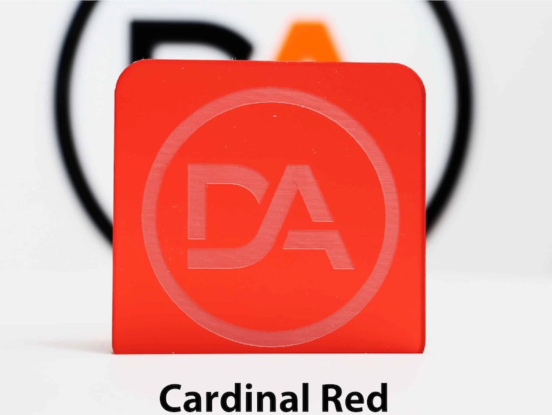 May include: A square, cardinal red object with rounded corners. The object has a circular, etched design with the letters "DA" inside. The text "Cardinal Red" is printed in a black, sans-serif font at the bottom of the image.
