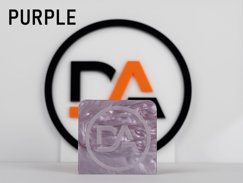 May include: A close-up of a purple, marbled, square-shaped sheet of acrylic with a white circle and the letters "DA" inside the circle. The sheet is sitting in front of a black circle with a white "DA" inside.
