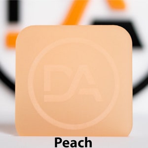 May include: A square, peach-coloured object with rounded corners. It features a circular logo with the letters "DA" embossed. The word "Peach" is printed in bold, black text at the bottom of the image.