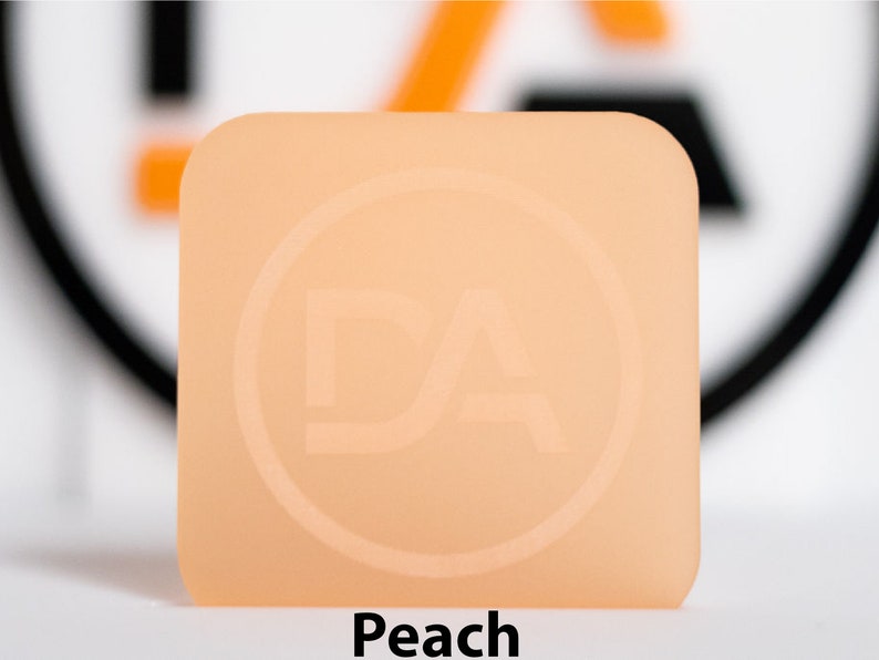 May include: A square, peach-coloured object with rounded corners. It features a circular logo with the letters "DA" embossed. The word "Peach" is printed in bold, black text at the bottom of the image.
