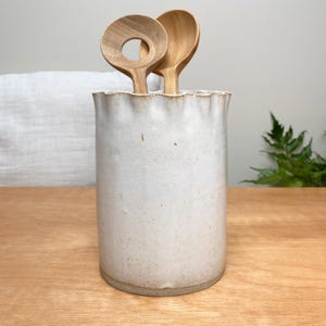 May include: A cream-coloured ceramic utensil holder with a wavy rim, containing two wooden spoons. The spoons are light brown, one with a circular hole. The holder is on a wooden surface.