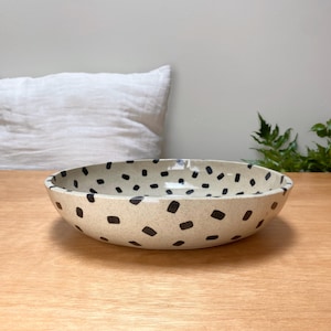 May include: A ceramic bowl with a speckled beige base and dark brown rectangular spots. The bowl has a wide, shallow design and sits on a wooden surface. The background includes a white pillow and greenery.