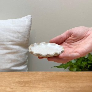 May include: A small, white ceramic dish with a scalloped edge. The dish is slightly oval in shape and has a speckled glaze.