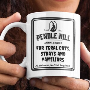 May include: White ceramic mug featuring the text "PENDLE HILL ANIMAL SHELTER" with a cat silhouette logo. The mug's design includes the phrases "FOR FERAL CATS, STRAYS AND FAMILIARS" and "All Welcome, No Trial Required".