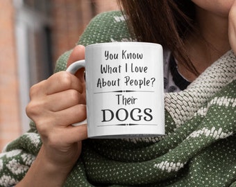May include: White ceramic coffee mug with black text that reads "You Know What I Love About People? Their Dogs". A person is holding the mug with their right hand.