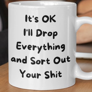 May include: White ceramic mug with a curved handle. The mug features black text that reads "It's OK I'll Drop Everything and Sort Out Your Shit". The mug is sitting on a wooden surface.