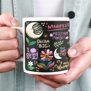 Op de afbeelding: Witte keramische mok met een zwarte achtergrond en kleurrijke illustraties. De tekst op de mok luidt "MANIFEST", "POSITIVE MINDSET", "DREAM BIG", "I AM STRONG", "YOU GOT THIS", en "I AM HAPPY".