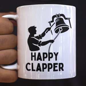 May include: White ceramic mug featuring a black graphic of a person ringing a bell, with the text "HAPPY CLAPPER". The mug has a curved handle. The mug is held by a person.