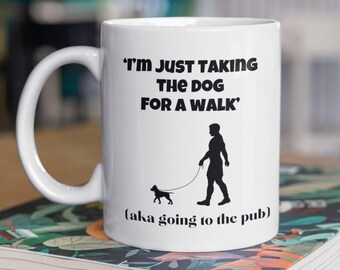 May include: White ceramic mug with a black silhouette of a person walking a dog. The mug has the text "I'm just taking the dog for a walk" and "(aka going to the pub)".