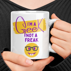 May include: White ceramic mug with a yellow and purple design. The mug features the text "I'm a Geek not a Freak" and a cartoon smiley face wearing glasses.