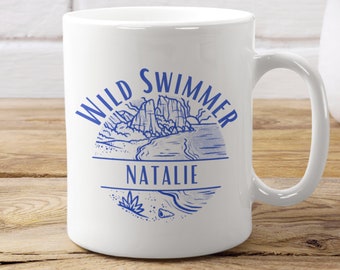 May include: White ceramic mug with a blue design of a mountain range, a beach, and the words "Wild Swimmer" with the name "Natalie" in the centre.
