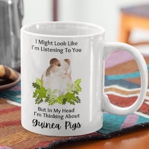 May include: White ceramic coffee mug with a black and white illustration of a guinea pig surrounded by green leaves. The text on the mug reads "I Might Look Like I'm Listening To You But In My Head I'm Thinking About Guinea Pigs."