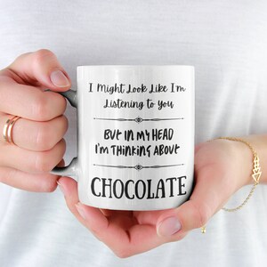 May include: White ceramic mug with the text "I Might Look Like I'm Listening to you BUT IN MY HEAD I'M THINKING ABOUT CHOCOLATE" in black. The mug is held by a person.