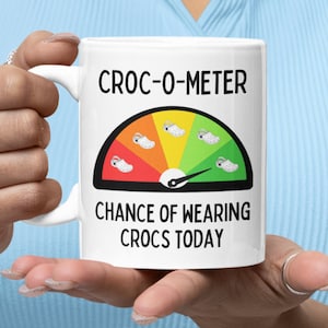 Op de afbeelding: Witte keramische mok met een zwart-witte afbeelding van een snelheidsmeter met de tekst "CROC-O-METER" en "CHANCE OF WEARING CROCS TODAY".