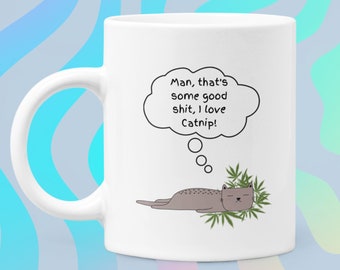 May include: White ceramic mug with a cartoon illustration of a gray cat lounging on catnip. A thought bubble above the cat reads, "Man, that's some good shit, I love Catnip!" The mug has a white handle.