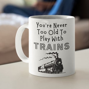 May include: White ceramic coffee mug with a black and white illustration of a train and the text "You're Never Too Old To Play With TRAINS".