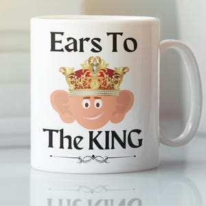 11oz ceramic mug featuring a cartoon king charles with large ears, wearing an ornate gold crown with red and blue accents, smiling with a quirky expression.The text is 'Ears to the King'