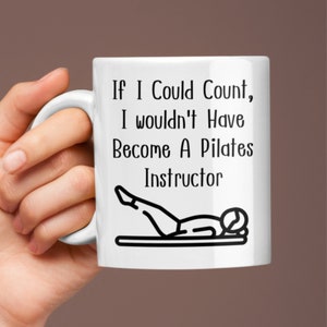 Puede incluir: Taza de cerámica blanca con un diseño negro que dice "Si pudiera contar, no me habría convertido en instructor de Pilates". El diseño también incluye una silueta negra de una persona haciendo un ejercicio de Pilates.