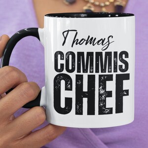May include: White ceramic mug with a black interior and handle. The mug features the text "Thomas COMMIS CHEF" in black, distressed font. The mug is held by a person.