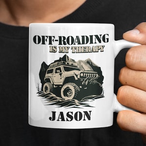 May include: White ceramic mug with a black and white illustration of a jeep off-roading. The text "Off-Roading is my therapy" is printed in black above the jeep and the name "Jason" is printed in black below the jeep.