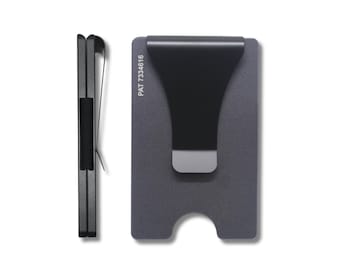 May include: A minimalist, gray metal card holder with a black money clip. The holder is rectangular with rounded corners and a curved cutout at the bottom. The text "PAT 7334616" is printed vertically on the front.